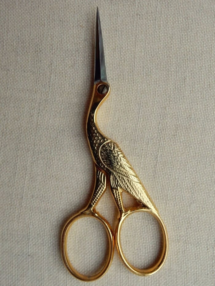 Golden bird-shaped embroidery scissors with a pointed tip on a beige fabric background.