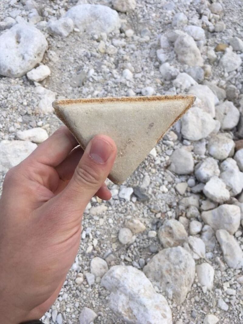 Hand holding a triangular stone artifact against a background of rocks and gravel.