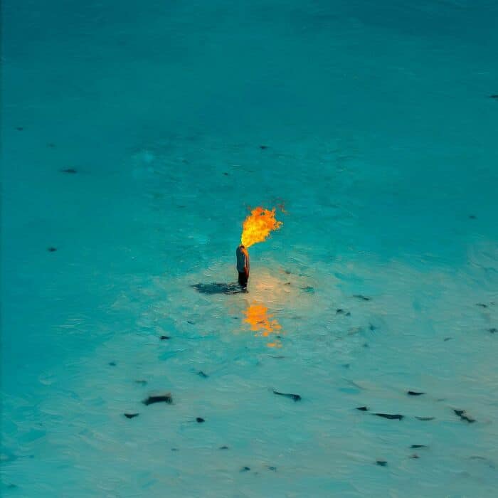 Performer breathing fire in a large, open blue landscape with a fiery reflection on the ground.