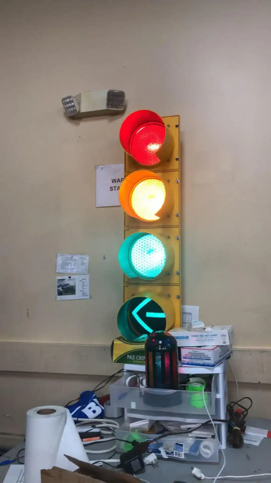 Traffic light with red, yellow, green, and arrow signals in a cluttered workspace.