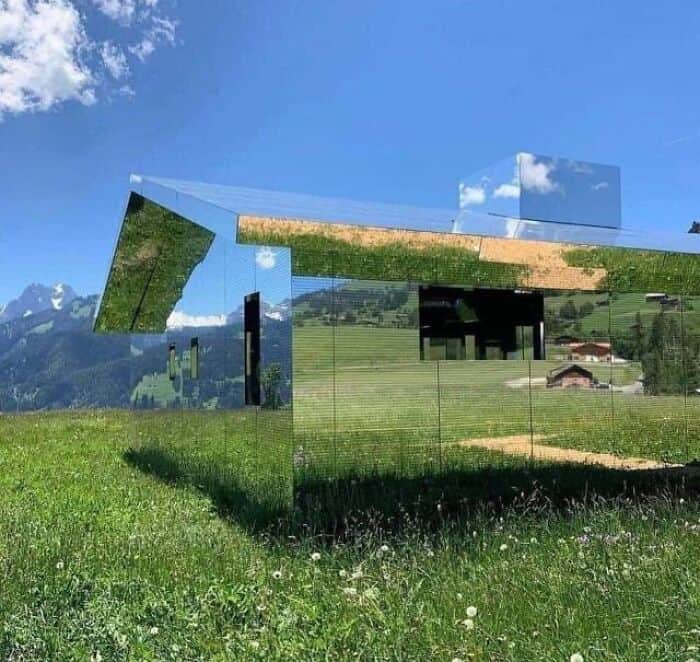 Reflective glass sculpture in a lush green field with mountains and blue sky.