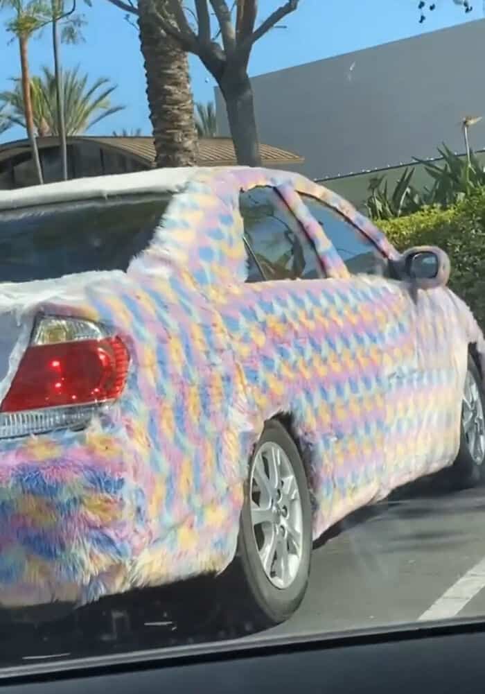 Car covered in multicolored fluffy fur wrap, parked outdoors on a sunny day.