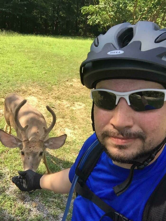 Cyclist wearing helmet and sunglasses taking a selfie with a deer in a green outdoor setting.