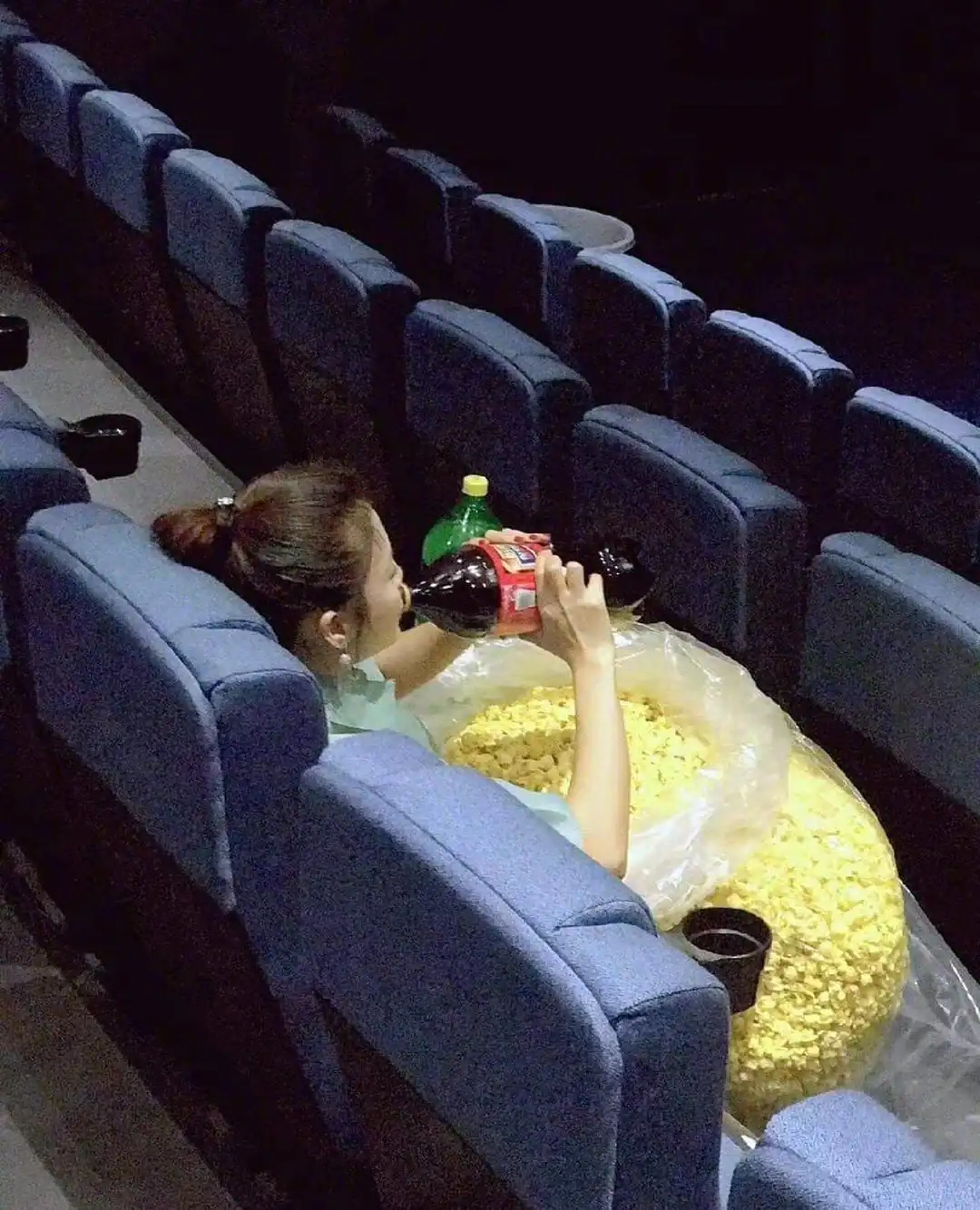 A woman sitting in a movie theater, drinking soda, with a large bag of popcorn in front of her.