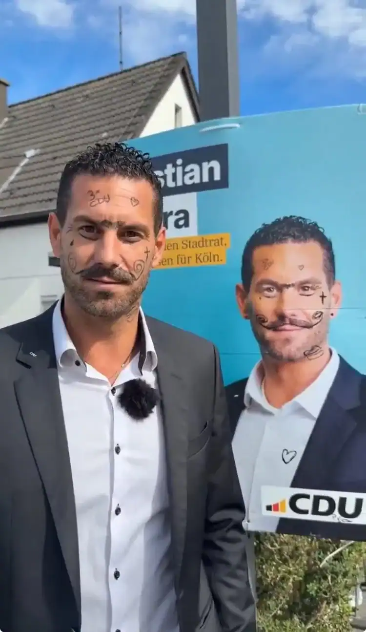 Man in a suit standing outdoors next to a political campaign poster with another man’s image.