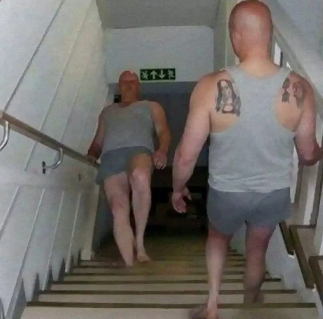 Two bald men walking up stairs, one with tattoos on his back, in a building corridor.