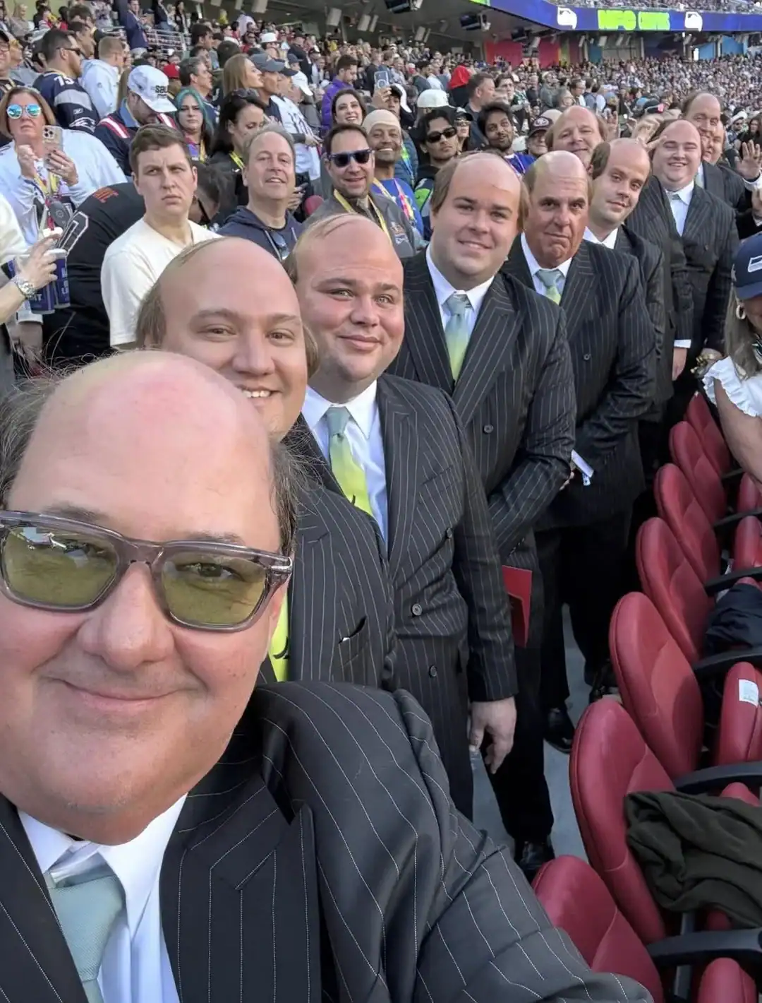 Group of men in suits at a stadium, smiling for a selfie.