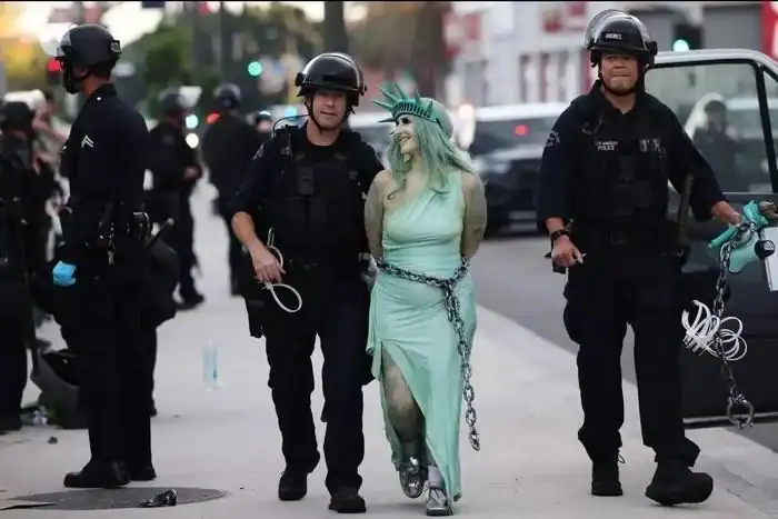 Police officers escort a person dressed as the Statue of Liberty, chained and in a light green dress.