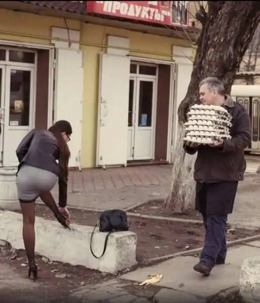 Man carrying a large cake in front of a building with a woman planting flowers on the sidewalk.