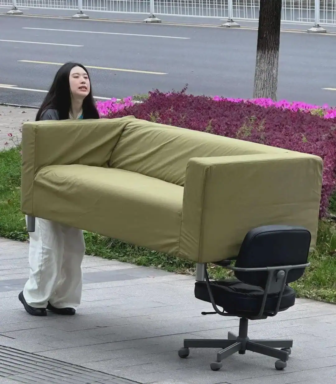 Woman carrying a large sofa on her back on sidewalk.