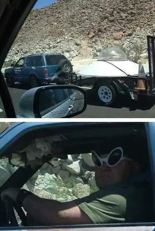 Man with sunglasses driving past a construction site with a trailer and a blue vehicle.