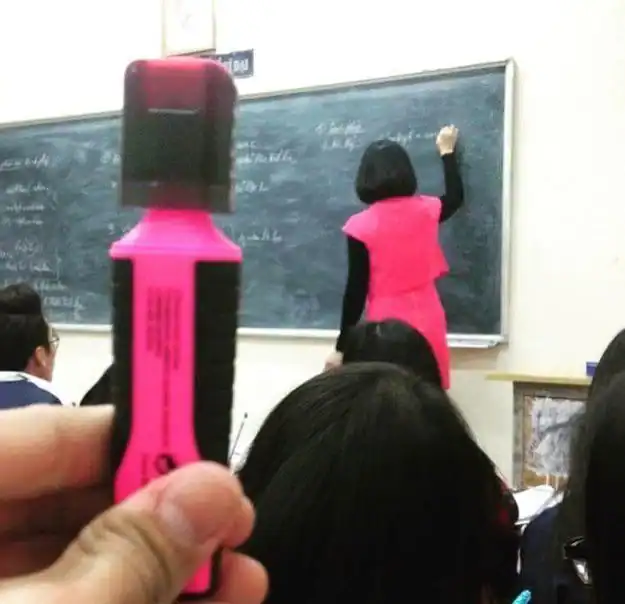 Teacher writing on blackboard in classroom with students, pink highlighter in foreground.