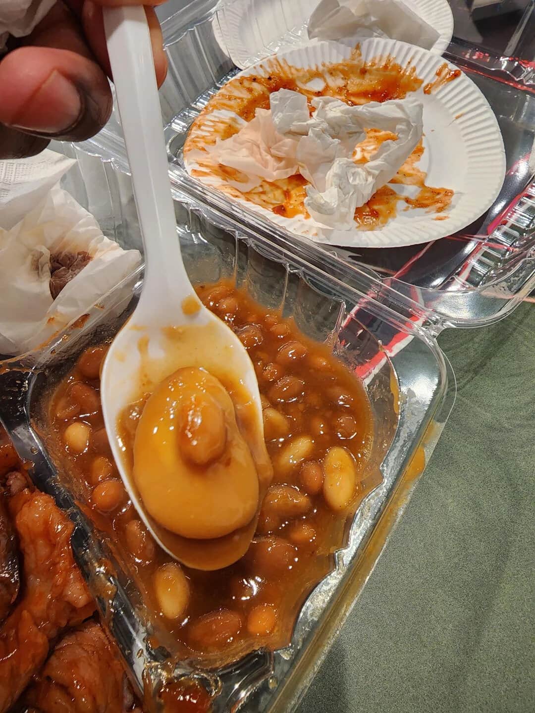 Spoon with egg and beans in sauce, paper plates with food in background.