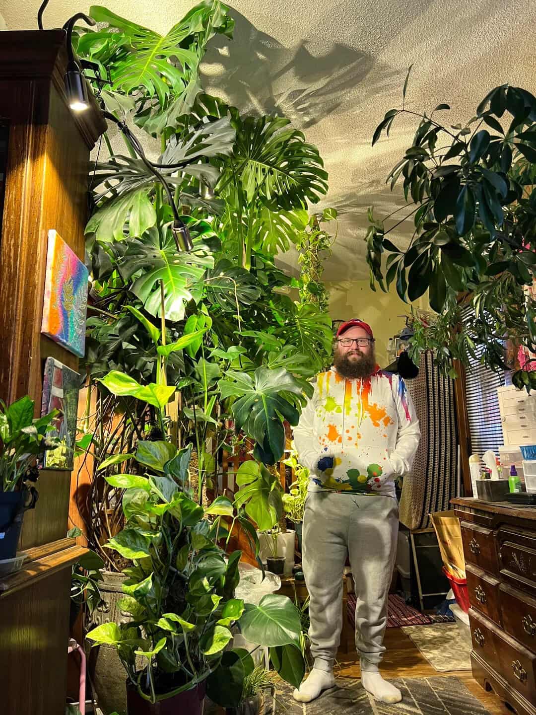 Man with a beard and glasses standing among large indoor plants in a cozy room.