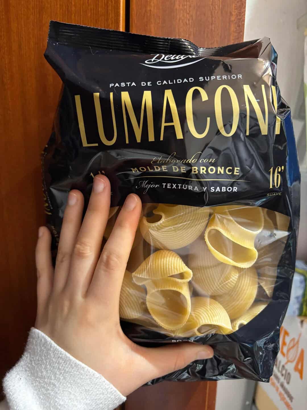 Hand holding a bag of Lumacon pasta with large shells, 16" size, against a wooden background.