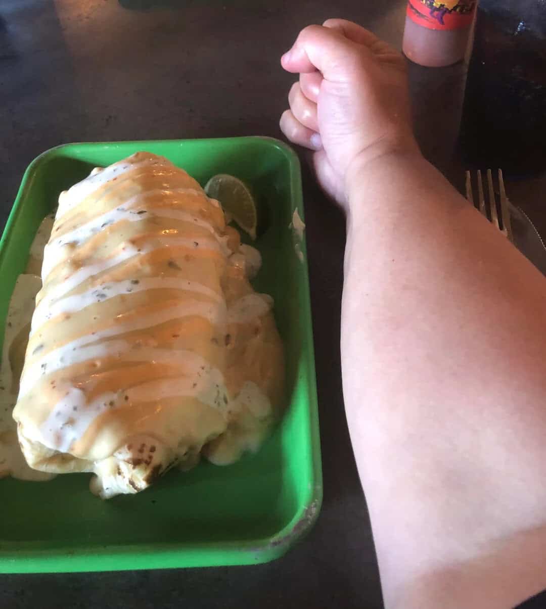 Large burrito with sauce on a green tray, arm and hand nearby, lime wedge inside.
