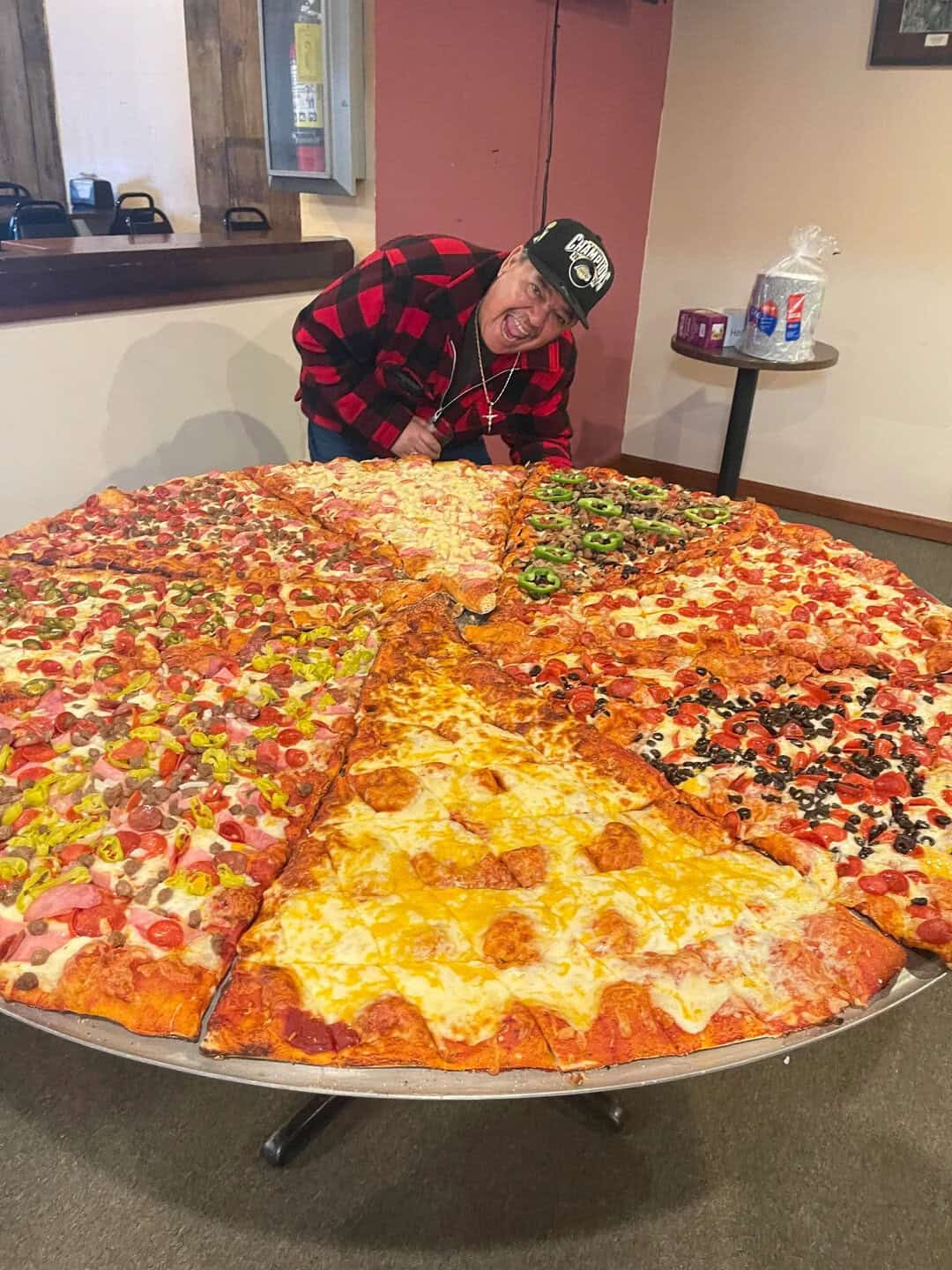 Person in a red and black checkered shirt leaning over a large pizza.