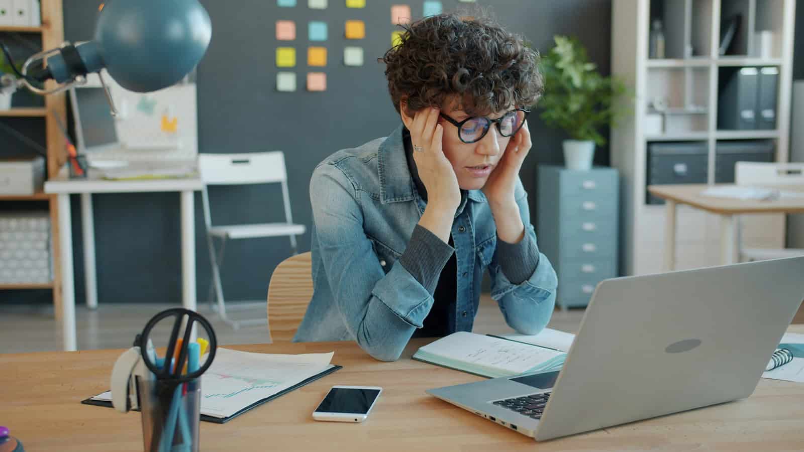 Frustrated woman with glasses at desk, holding head, working on laptop.