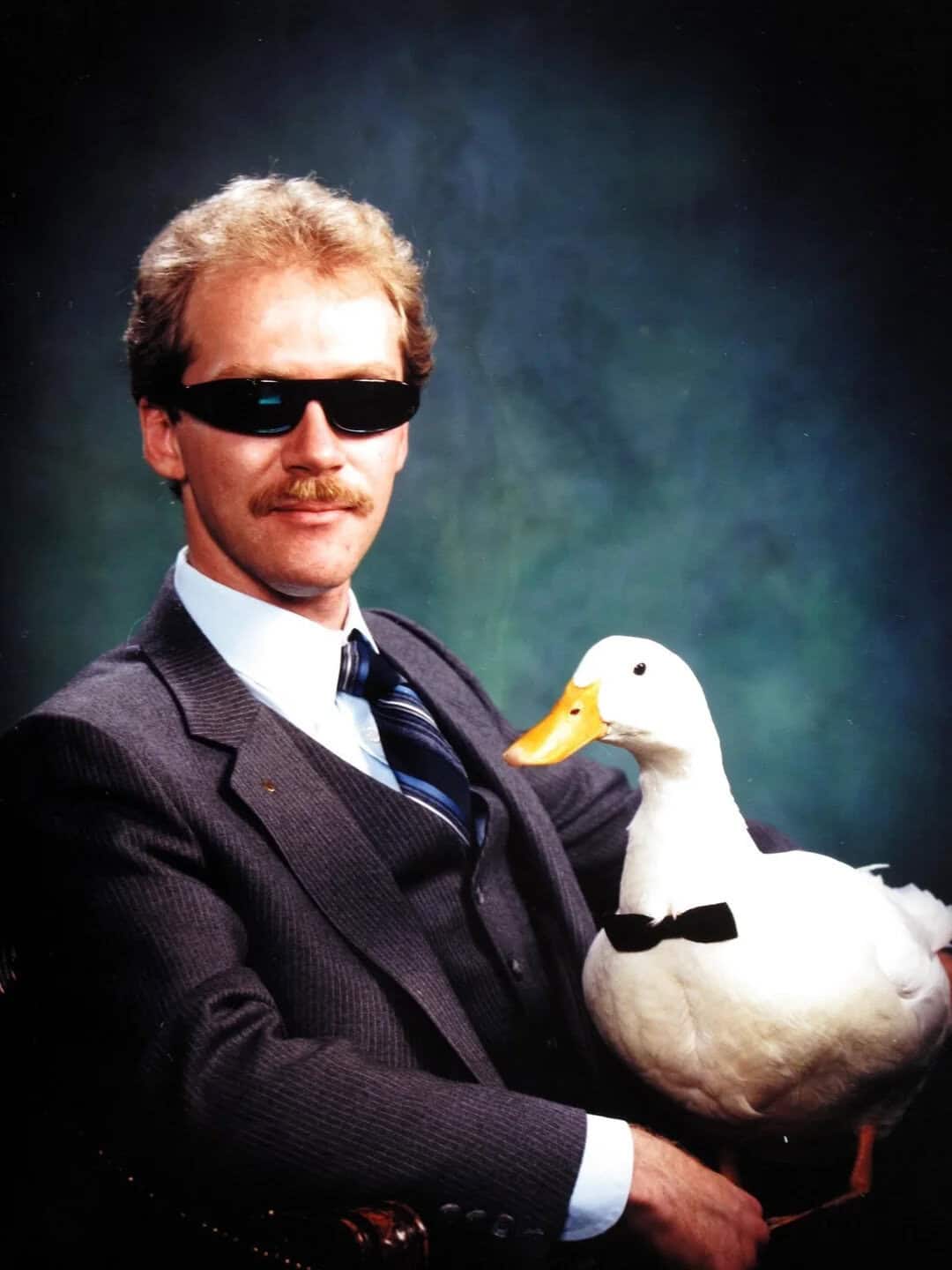 Man in suit with sunglasses holding a duck in a studio setting.