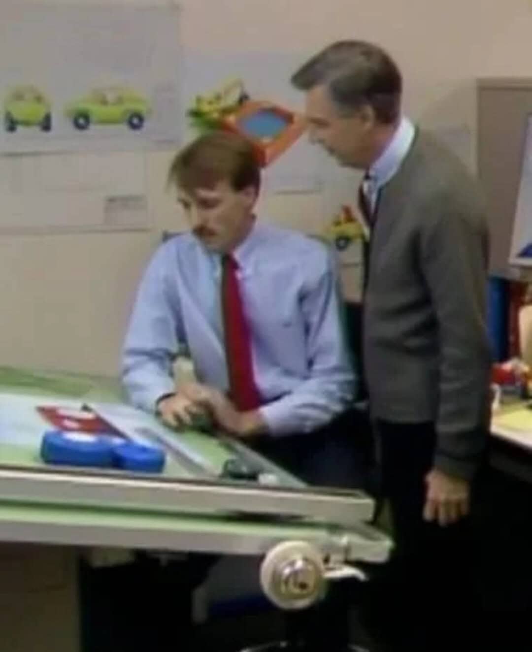Two men in an office, one sitting and one standing, looking at a model car on a desk.