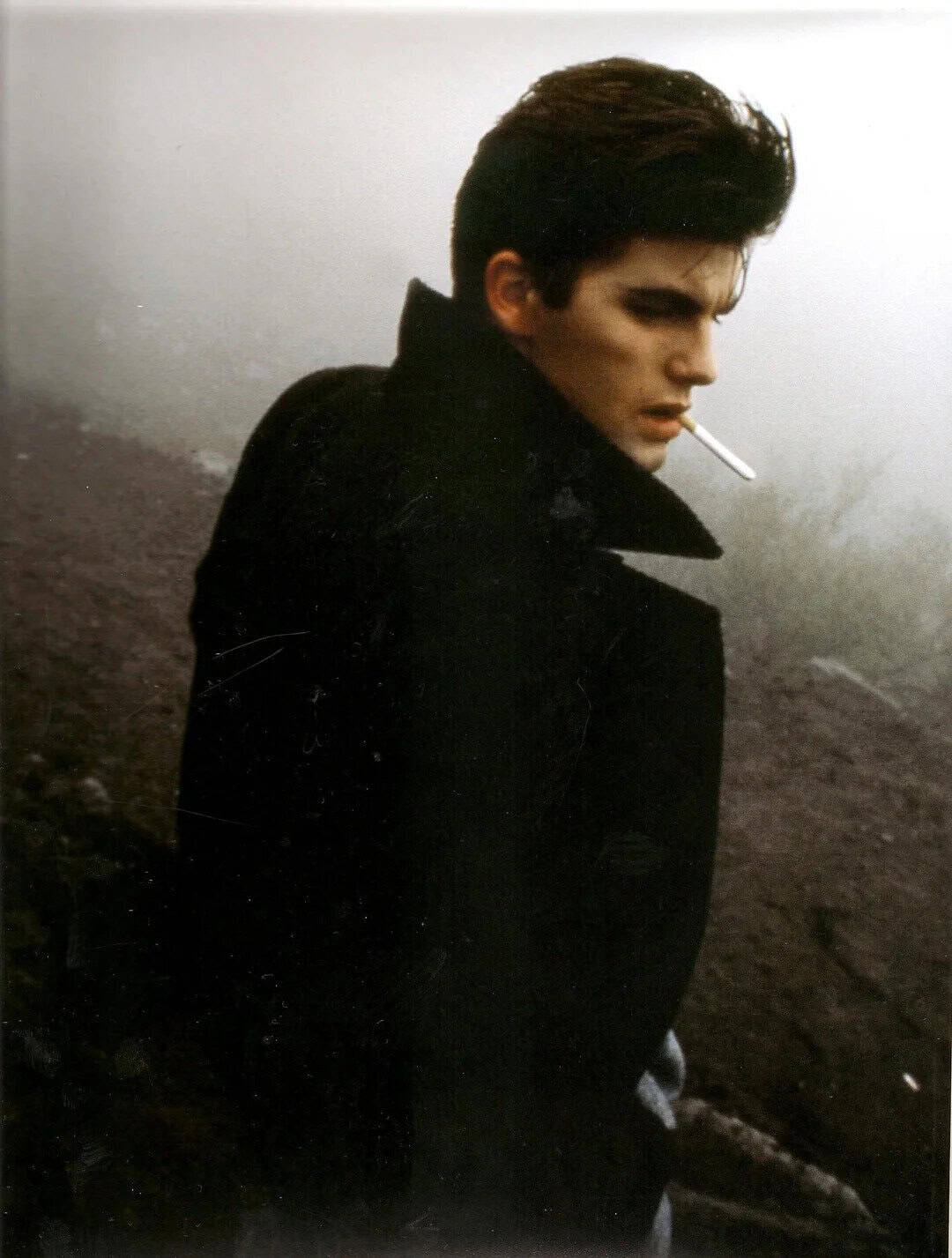 Young man with dark hair, smoking a cigarette, wearing a black coat, in a foggy outdoor setting.