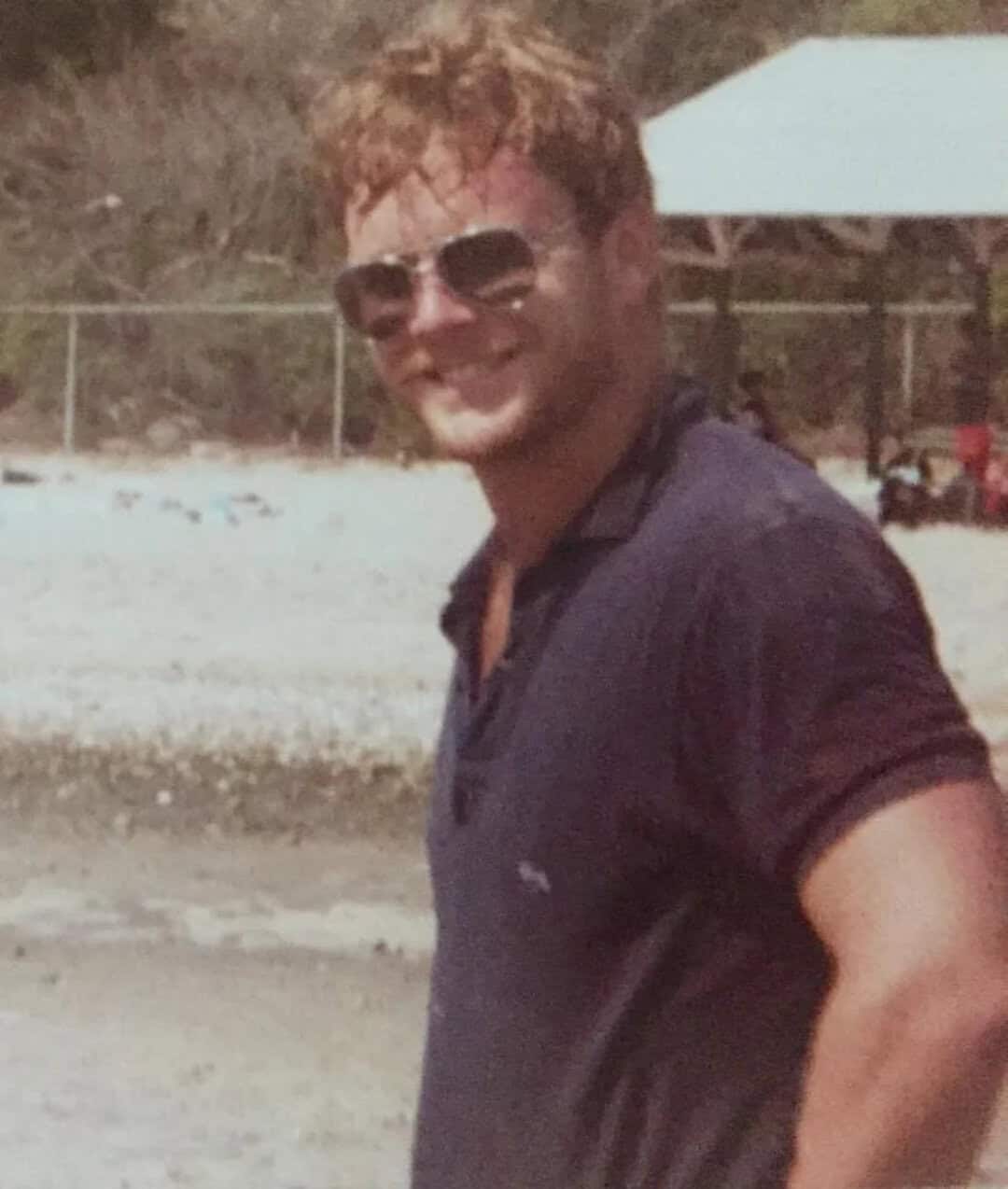 Young man with curly hair wearing sunglasses and a navy polo shirt, smiling outdoors.