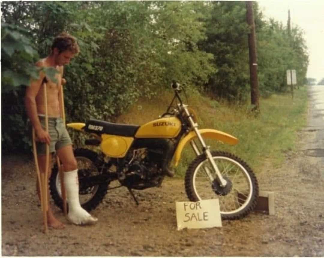 Young man with crutches and a cast on his leg standing next to a yellow Suzuki motorcycle for sale.