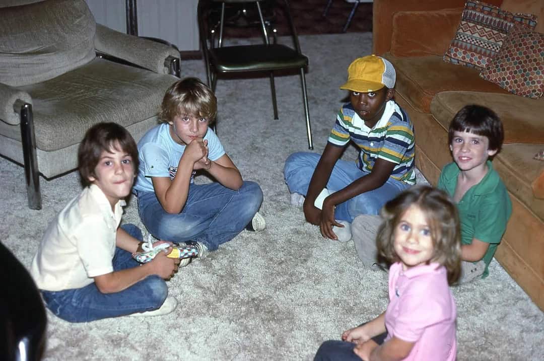 Group of children sitting on carpet in living room, one with a yellow cap.