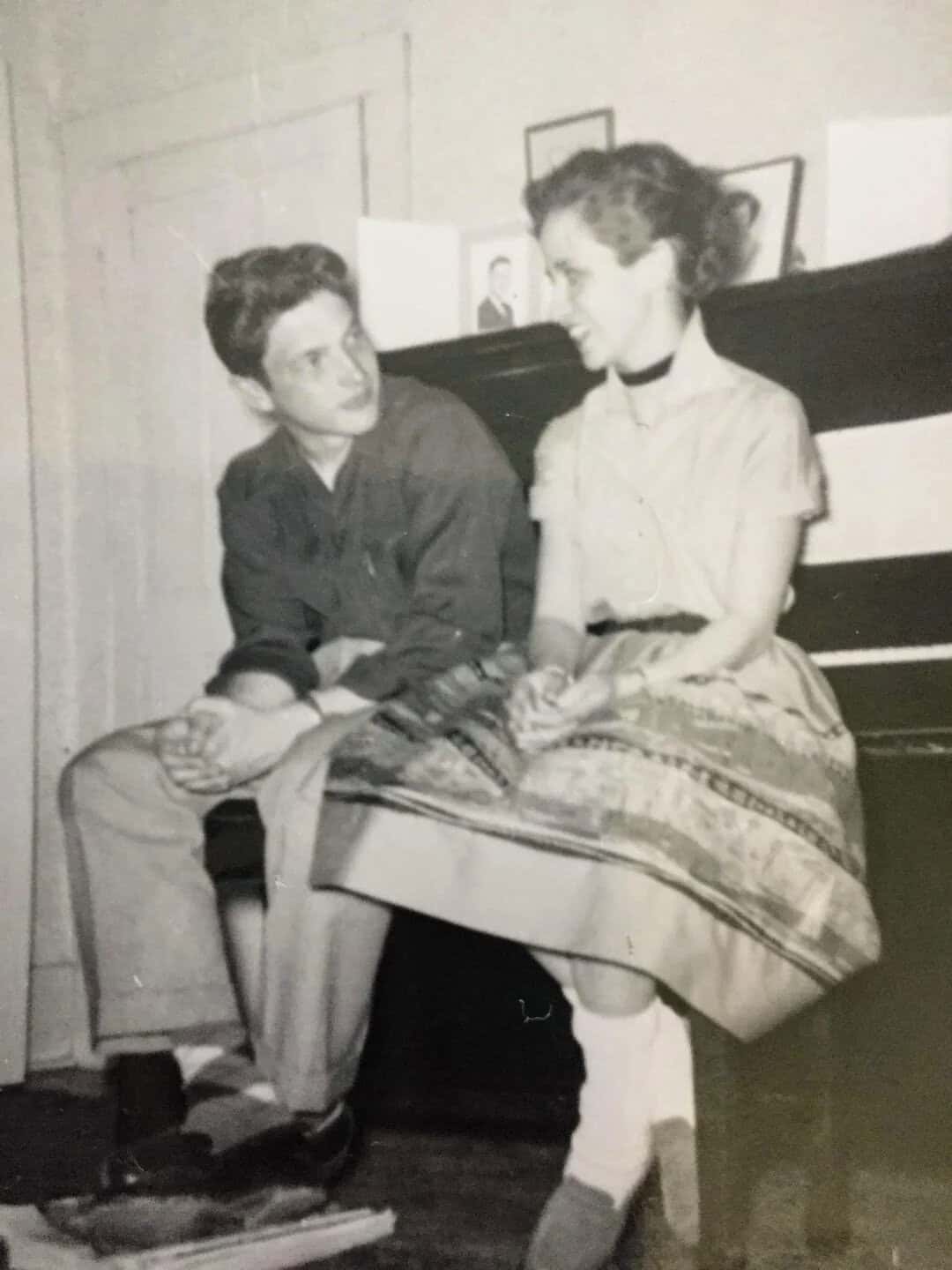 Black and white photo of a young man and woman sitting on a piano bench, talking.
