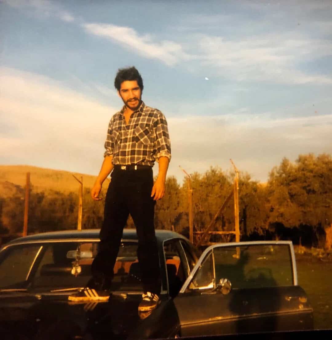 Man with beard in plaid shirt standing on a car in an open field.