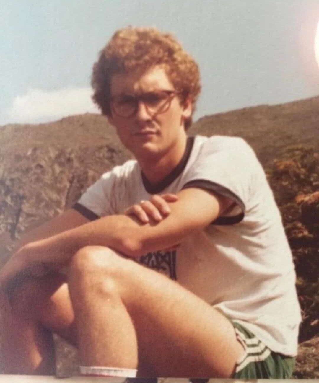 Young man with curly hair and glasses sitting outdoors on a hillside.