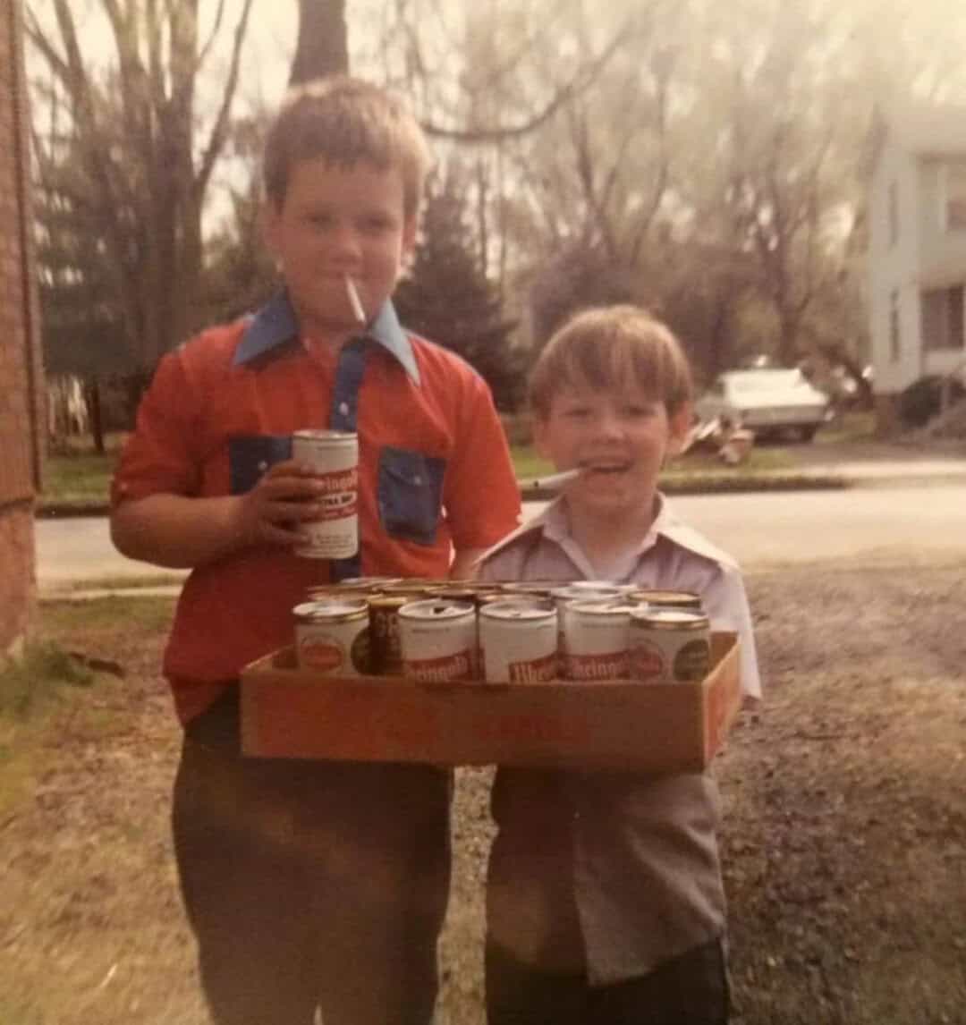 Two young boys with cans of beer, one holding a box of cans, outdoors in a suburban neighborhood.