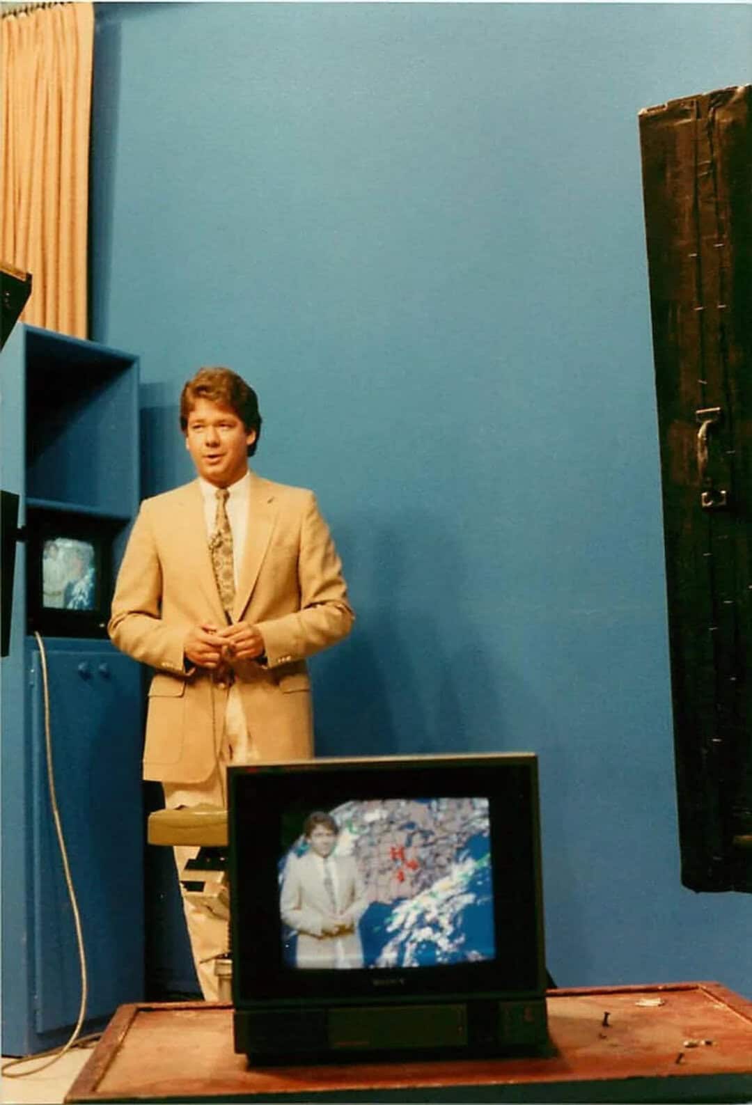 Young man in beige suit standing in front of blue wall with TV showing weather map.