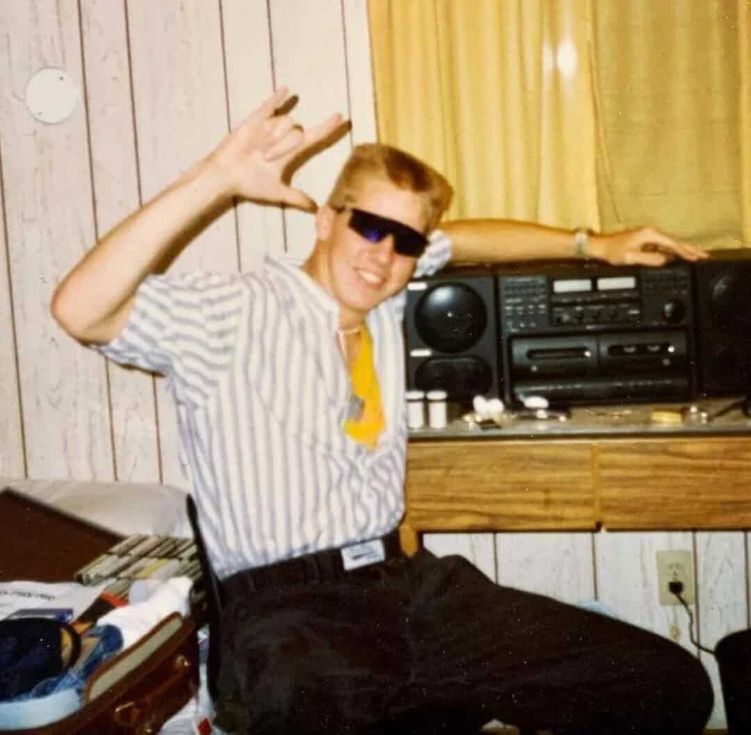 Young man with sunglasses, striped shirt, and yellow tie, waving and smiling.