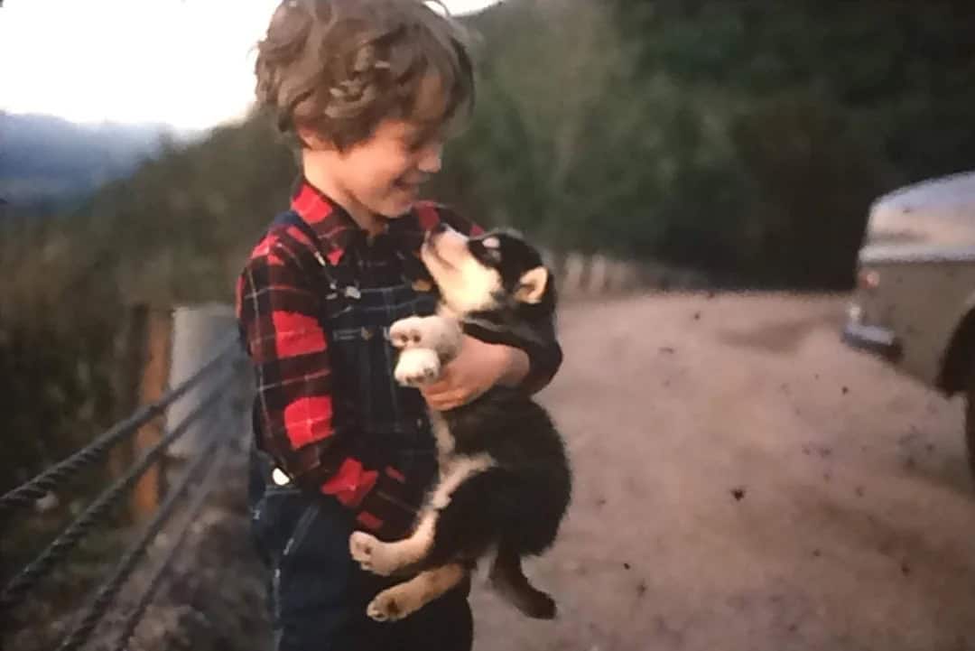 Child in red plaid shirt holding a puppy, smiling outdoors.