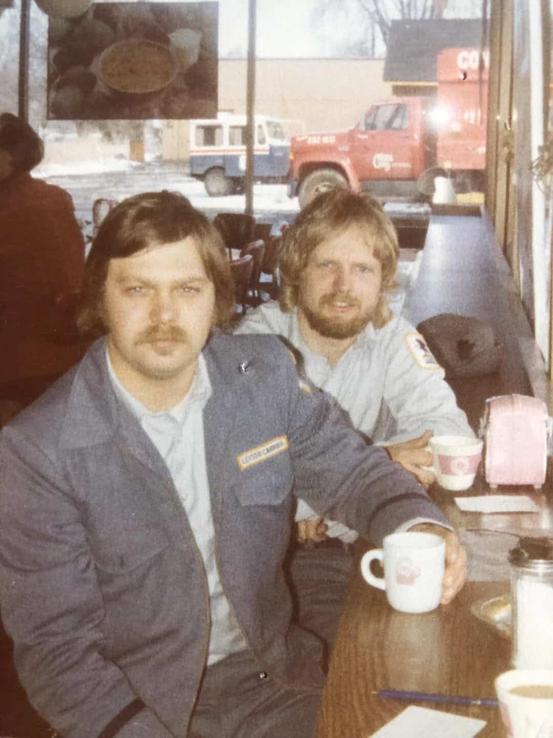 Two men with beards and long hair sitting at a table in a cafe, one holding a coffee mug.