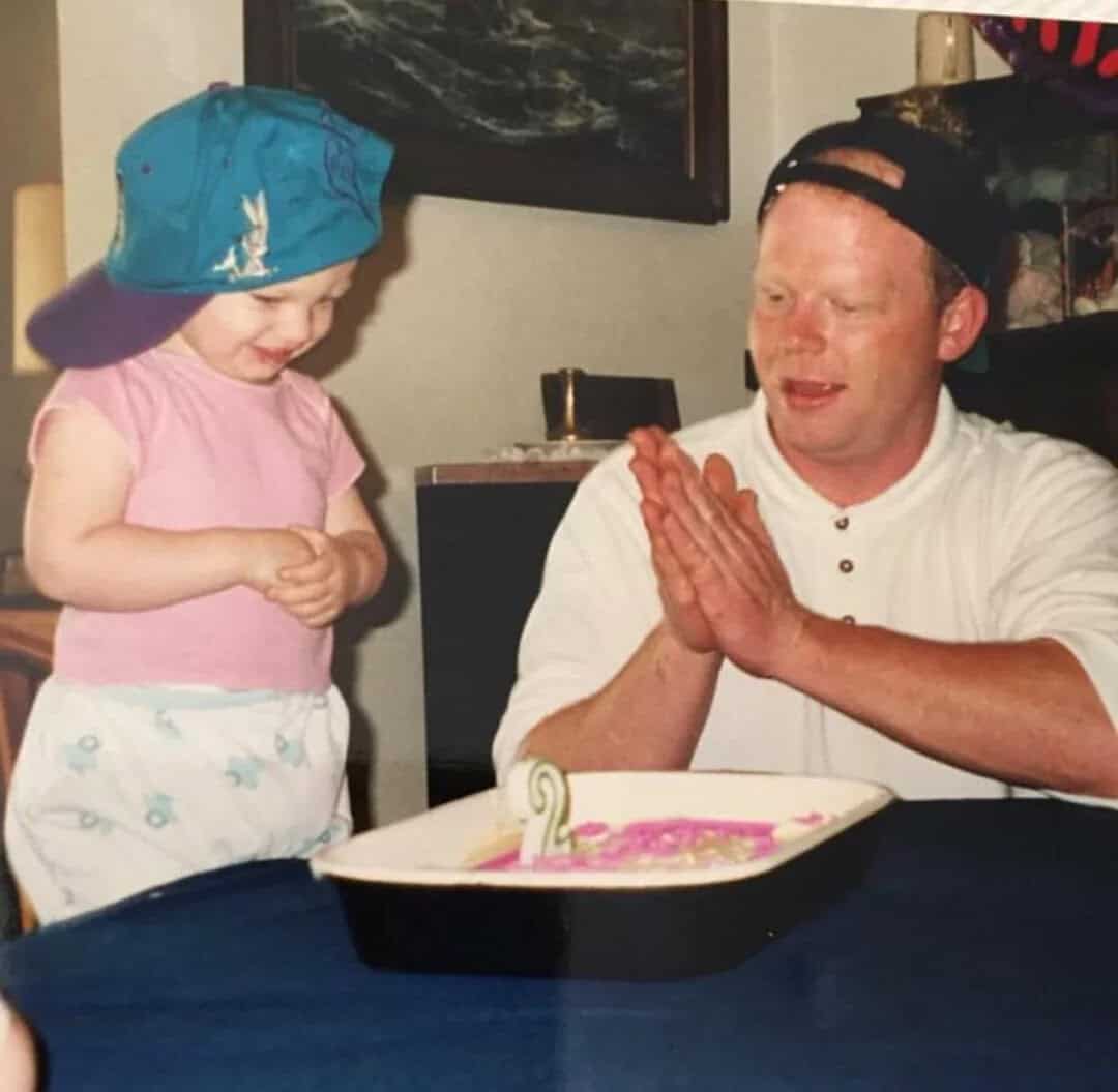 Father and daughter celebrating birthday with cake, girl wearing blue cap and pink shirt.