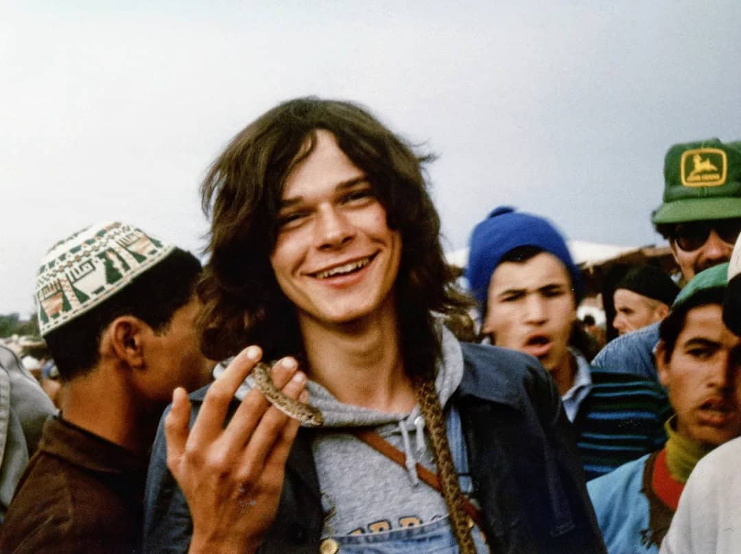 Young man with long hair smiling, holding a cigar, surrounded by people.