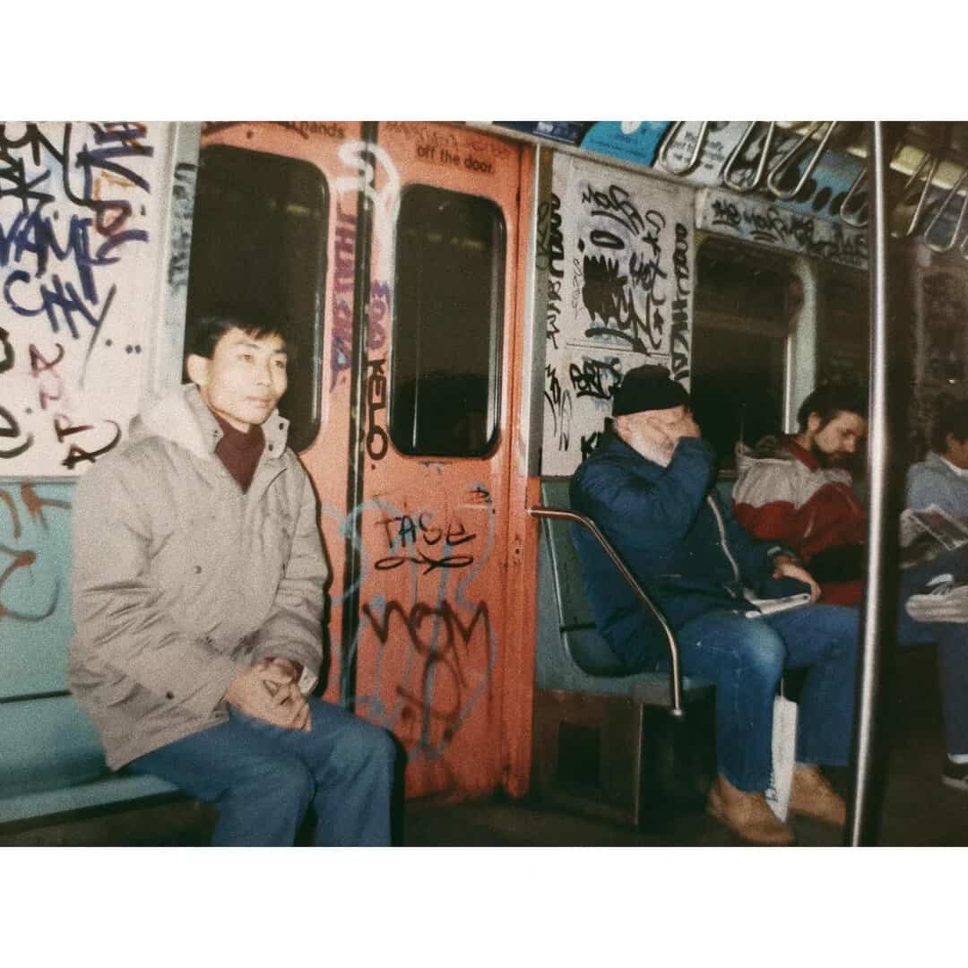 Young man sitting on subway with graffiti-covered walls, wearing a beige jacket.