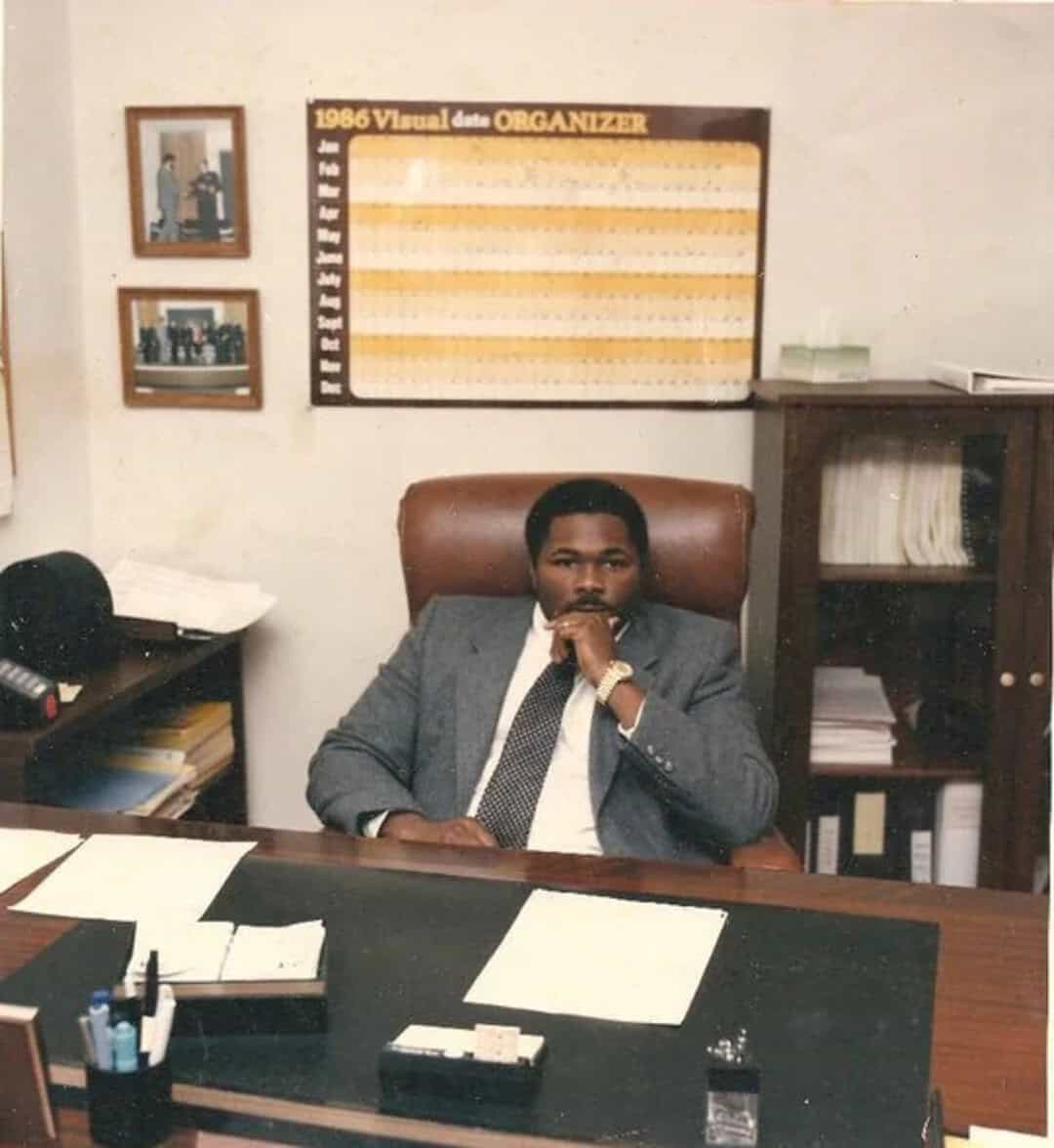 Man in suit sitting at desk in office with 1986 visual organizer on wall.