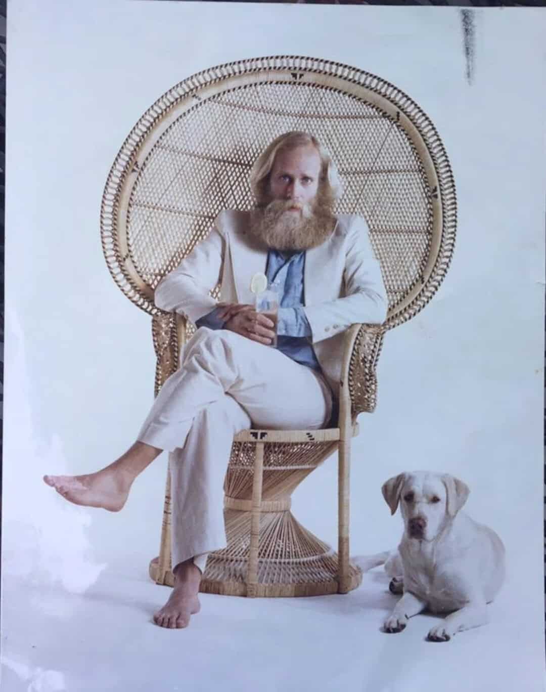 Man with long beard sitting on wicker chair with dog beside him.