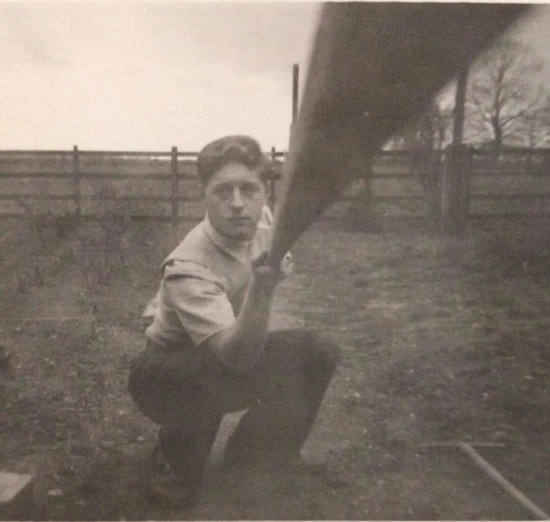 Young man crouching outdoors holding a long object, possibly a rifle, in black and white.
