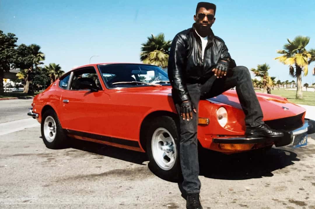 Man in black leather jacket and sunglasses leaning on red vintage car in sunny park.