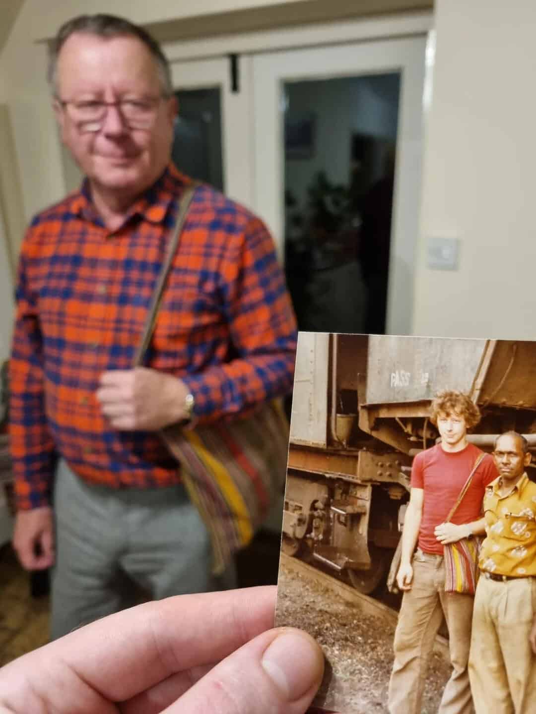 Older man in plaid shirt holding a striped bag, with a photo of two young men in front of a train.