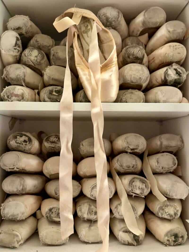 Ballet slippers hanging in front of a shelf of rolled fabrics or textiles.