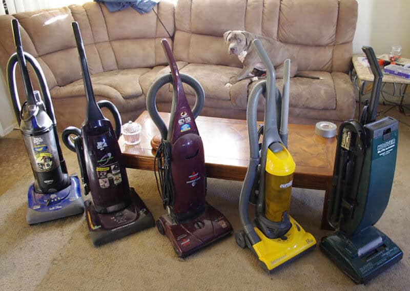 Collection of five vacuum cleaners in front of a beige sofa.