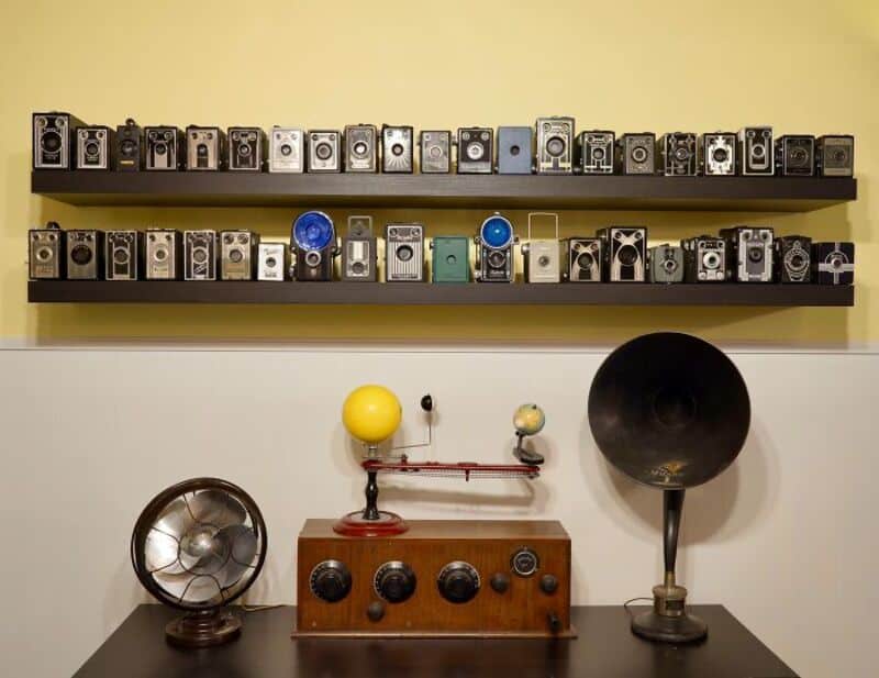 Collection of vintage cameras on shelves with a radio and gramophone on a table.