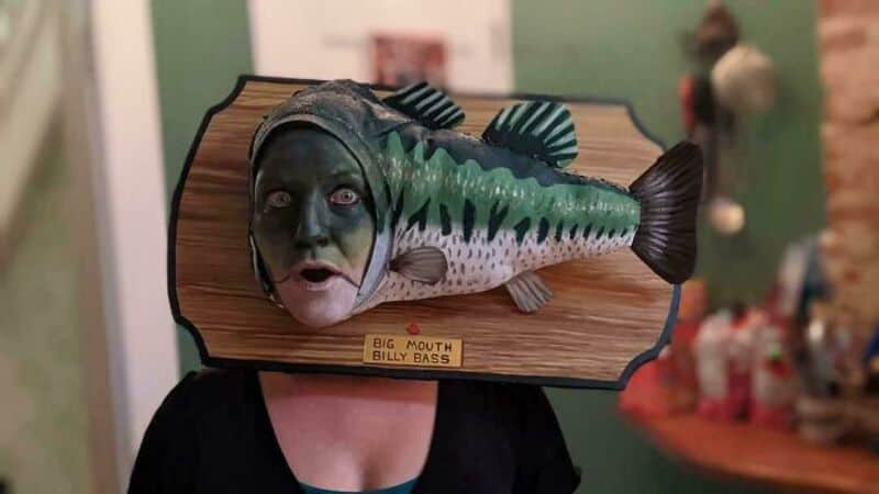 A large fish model with a human face mounted on a wooden plaque.