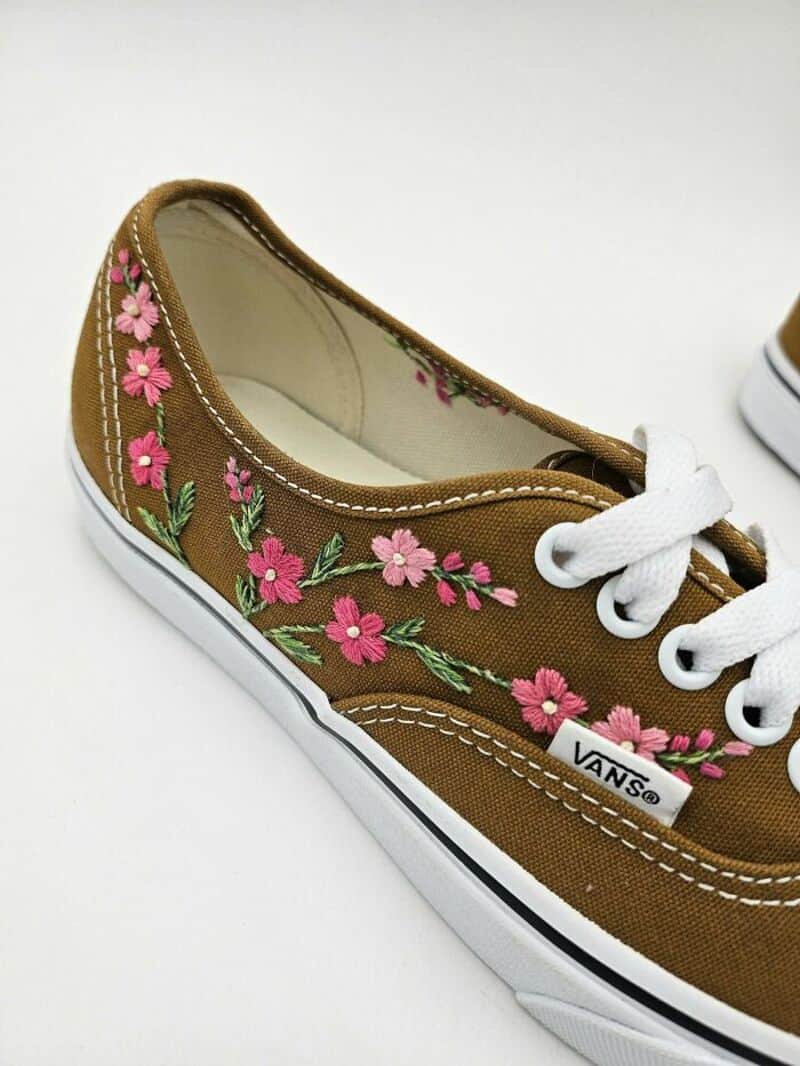 Vans sneaker with pink floral embroidery on brown canvas, white laces, and sole.
