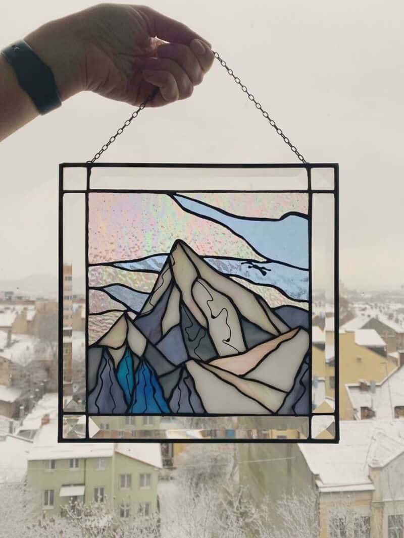 Stained glass window of mountain landscape held by hand against snowy city background.